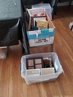 Cardboard box filled with assorted cookbooks and a Bible and a plastic tub containing various picture frames on a wooden floor near a table.