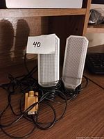 Two rectangular Dell speakers with light gray mesh fronts and white plastic edges, placed on wooden surface with connected cables tangled around them, including a yellow connector plug.