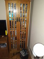 Front view of tall brown wooden cabinet with glass front doors showing DVDs inside and black metal handles, in good condition.