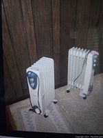 Photo showing two white oil-filled radiator electric heaters with caster wheels and thermostat control panels, placed on carpeted floor in front of wood-paneled wall.