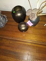 Three bronze decorative vases on a wooden surface, two round vases and one with dried floral stems including lotus seed pods.