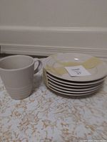 Stack of white ceramic plates and a white ceramic mug with handle on a textured surface in front of a white wall.