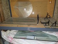 Photo of clear glass cabinet door, round glass piece, two flat glass shelf pieces, and two black decorative metal shelf brackets displayed on a gray table with pegboard wall background.