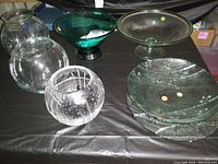 Five glass items including bowls and vases on black cloth: clear pitcher vase, spherical etched vase, large textured clear bowl, green pedestal bowl, and clear pedestal compote bowl.