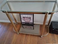 Gold tone metal stand with two glass shelves shown resting on wooden floor against a wall.