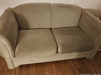 Beige fabric love seat sofa with two seat cushions and sewn tear visible on back corner.