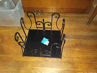Black metal magazine rack on wooden floor with decorative scrollwork and square base for holding magazines.