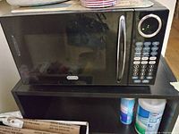 Black microwave oven with digital keypad and handle on a shelf, with cleaning supplies underneath.