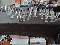 Photo shows multiple clear glass items on a black tablecloth including water pitcher, multiple wine glasses, salt and pepper shakers with floral decorations, and various drinking glasses or canisters