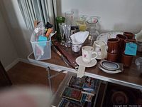 Image shows a collection of kitchen items on a folding table including brown coffee cups, clear glass vases, white measuring cups, white ceramic mugs with designs, and various utensils and containers.
