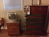 Photo of the wooden night stand with two drawers and a vintage television on top next to a tall wooden dresser wrapped with green straps around the drawers, in a room with window blinds.
