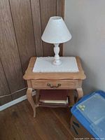 Photo showing a wooden night stand with a drawer, curved legs, lower shelf with folded items, and a white table lamp with a fabric shade on top, placed on a crocheted doily.