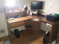 L-shaped office desk in light brown wood finish. Features under-counter keyboard tray, one drawer, raised monitor shelf with flat screen monitor. Desk is used and needs disassembly.