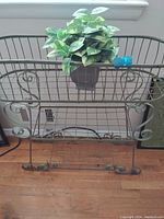 Green metal plant stand with decorative scrollwork, basket-style top, and bottom shelf. Displaying a potted vine plant on the top basket.