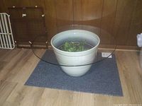 Round clear glass table top displayed on a white flower pot base for size reference. The setting is an indoor room with wood panel walls and hardwood floor.