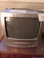 Photo showing a silver CRT TV with curved screen on wooden surface, visible front buttons and input ports, used condition.