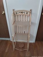 Tan decorative kitchen stand with three wire shelves and floral design on the top back panel, standing against a wall between two doors.