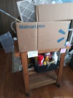 Wooden utility cart with two shelves and wheels, holding a cardboard box and a basket of cleaning supplies and hardware items.
