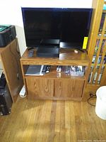 Wood TV stand with flat screen TV, DVD player, remotes, open shelf, and two closed cabinet doors below. Medium brown wood grain finish. Used condition with minor wear.