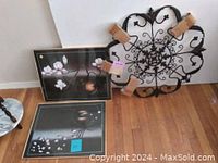 Photo showing one black metal ornamental wall plaque with scrollwork and two black framed pictures with floral images.