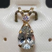 Front view of yellow gold pendant showcasing pear-shaped aquamarine and small round clear accent stone in prong settings against white display background.