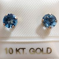 Photo showing close-up of two round blue topaz gemstones set in 10K yellow gold prong settings with a label stating 10 KT GOLD.