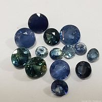 Top-down photo showing the assortment of 13 round faceted blue sapphire and related gemstones on a white background with colors ranging from deep blue to teal.