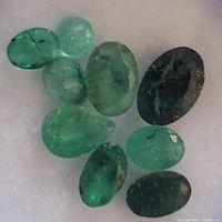 Close-up photo showing eight loose faceted emerald gemstones arranged on a white background with various oval and pear shapes, medium to dark green.