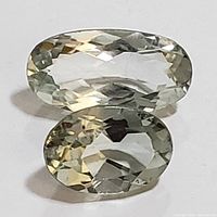 Top view of two oval faceted green amethyst gemstones placed on a white surface showing clarity, cut and light color.