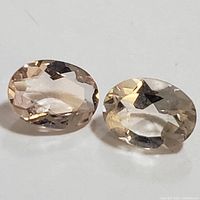 Close-up photo of two oval cut Morganite gemstones with light peach and pale pink colors side by side on white surface.