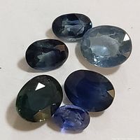Photo of six loose oval cut sapphire gemstones in varying blues and greenish hues on white background.