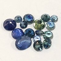 Loose genuine sapphire gemstones shown from above, various shapes and sizes, displaying blue and green hues