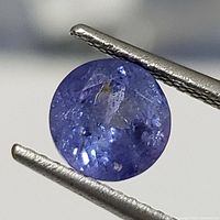 Close-up image of a round cut genuine Tanzanite gemstone held by tweezers showing natural inclusions and color saturation.