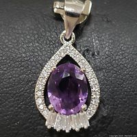 Close-up photo of the pendant showing the amethyst center stone, round cubic zirconia border, and baguette-cut cubic zirconia accents against black background.