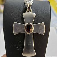 Close-up image of polished sterling silver cross pendant with oval garnet gemstone center set, mounted on black display stand.
