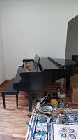 Side view of Baldwin black baby grand piano with keyboard cover open showing interior strings and hammers. Piano bench visible to left and household items on floor.