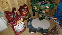 Two large sealed bags of Scott's GrubEx and green plastic storage seat with beige lid, wheeled gardening spreader seen behind them.