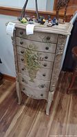 Front view of the shabby chic aged white hand-painted jewelry armoire with floral vase artwork on drawers and small decorative items on top.