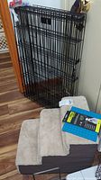 Fold-up black metal wire pet crate folded and leaning against wall, beige carpeted pet step-up assist with three steps on floor, blue puppy lickimat package on top of step-up assist.