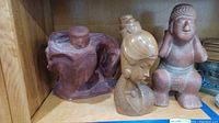 Wide shot showing three figural sculptures on a wooden shelf: abstract reddish-brown sculpture, beige bust of a woman, and a seated reddish-brown figure sculpture.