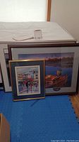 Group view of two framed and matted prints leaning against a wall on a blue floor mat.