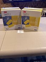 Two boxes of 3M TaperFit 2 ear plugs sitting on a table, showing front packaging with branding and yellow ear plugs graphic.