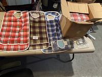 Multiple adult bibs in different plaid patterns laid flat on a table with some packed in a cardboard box.