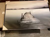 Photo showing the full framed print featuring the wooden pier scene, with protective corners on the frame.