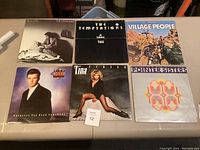Six vinyl record albums laid out on a surface, showing front covers by Billy Joel, The Temptations, Village People, Rick Astley, Tina Turner, Pointer Sisters.