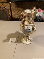 Front and angled side views of a decorative Italian pottery vase with gold-toned scroll handles, colored stones or glass adornments in blue, red, and turquoise, and an ornate pedestal base with gold accents.