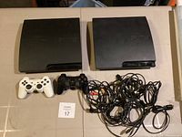 Two black PS3 Slim consoles with white and black DualShock 3 controllers and assorted cables laid out on a flat surface.