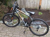 Side view of Avigo Mountain Series Ultra 26 mountain bike showing metallic gray frame with orange branding, front suspension fork, knobby tires, black seat, and black pedals standing on a stone-paved ground outdoors near plants.