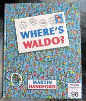 Front cover of the first edition Where's Waldo? book showing detailed crowd illustration with Waldo hidden and title in a postage-stamp style frame.