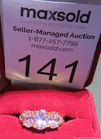 Front view of rose quartz ring showing three oval pink stones and surrounding small clear stones on a gold-tone band in red ring box